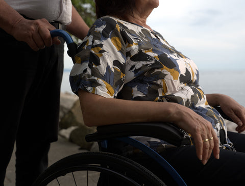 A woman sits in her blue Strongback Comfort Flip wheelchair on a waterside path at dusk while a companion stands behind with a hand on the push handle