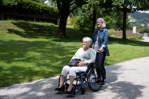 Caregiver pushing an older woman seated in a blue Strongback Comfort S wheelchair along a park path