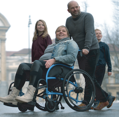 A woman sitting upright in a blue Strongback Heavy Duty wheelchair is pushed along a city street by a man, with two children walking alongside