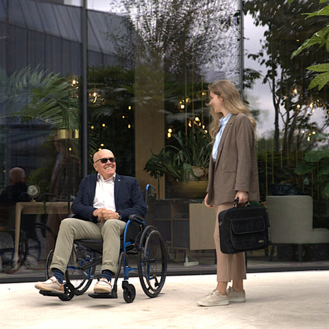 A man sits upright in his blue Strongback Comfort wheelchair outside a glass-fronted cafe, talking with a woman standing beside him