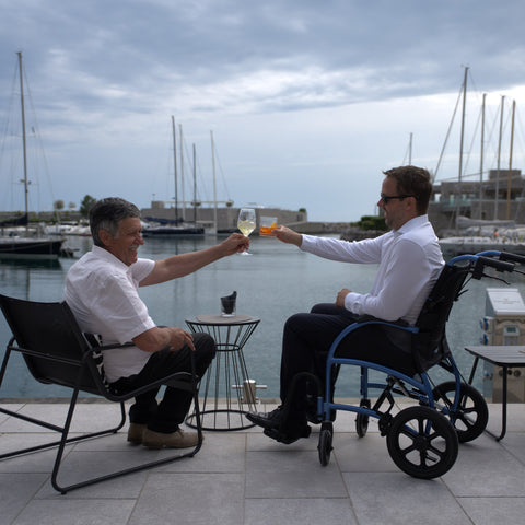 A man seated in his blue Strongback Excursion transport chair raises a glass with a friend at a marina terrace table
