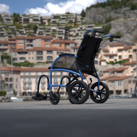 Strongback Excursion transport chair from the side on a marina boardwalk, showing the 12 inch rear wheel with no hand rim and the attendant brake lever on the push handle